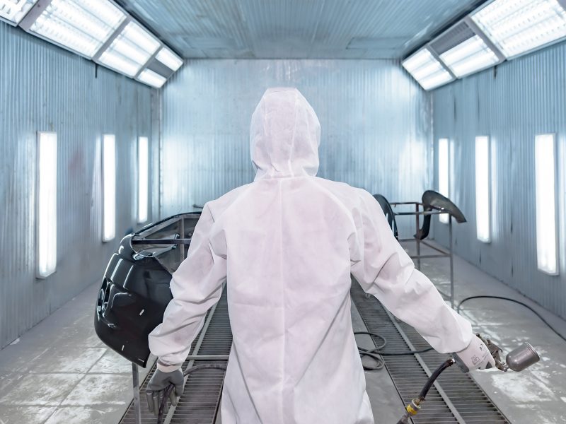 automobile repairman painter in protective workwear and respirator painting car body in paint chamber